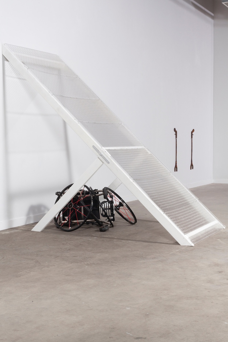 A larger than life ramp is positioned at a steep angle attaching the white wall of the gallery space to the grey floor. Underneath the ramp is a broken black wheelchair. In the distance hanging on the wall is an artwork titled *Grabbers*, consisting of two grabbers similar to the ones Emily uses.