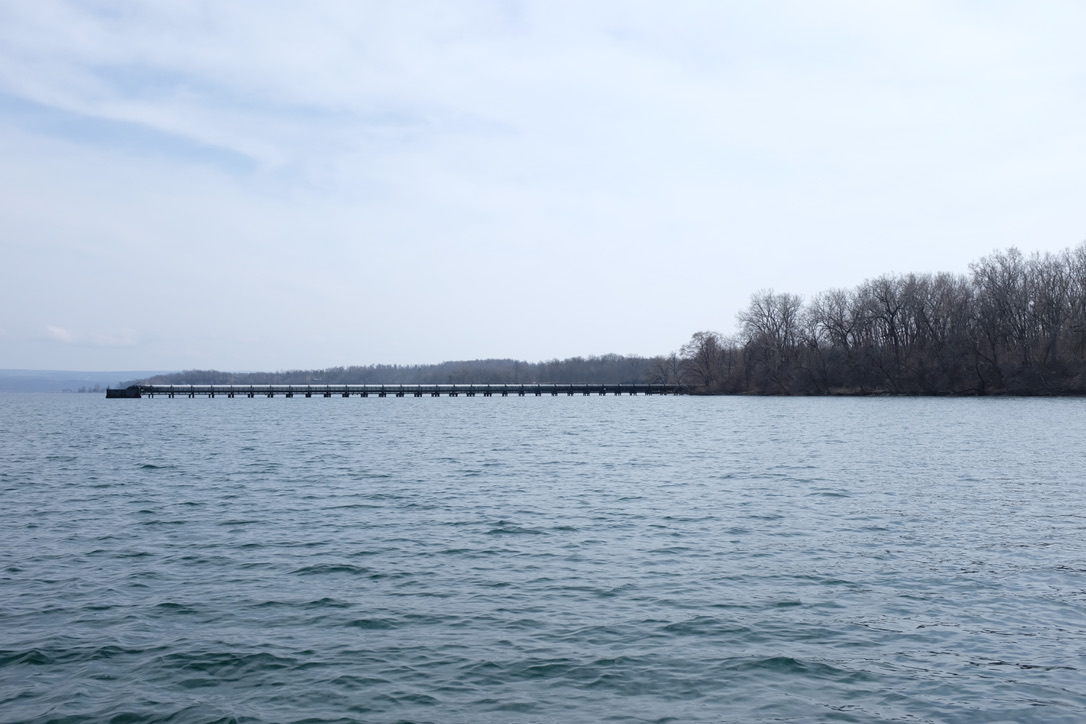 a wide shot of a long metal pipe extending from a woodsy shore into the waters of Seneca Lake